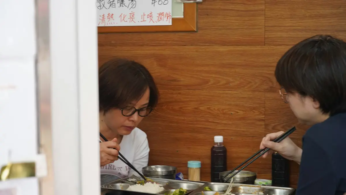 兩位女士在香港地道小食店內用餐 ,以筷子進食,背景可見中文字招牌及典型香港茶餐廳裝潢,場景真實自然,極具香港本地特色。